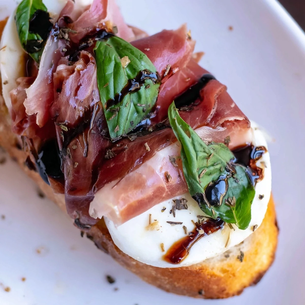 Golden toasted baguette slices topped with melted mozzarella, roast beef, and prosciutto, finished with fresh basil and balsamic glaze on a rustic platter.  