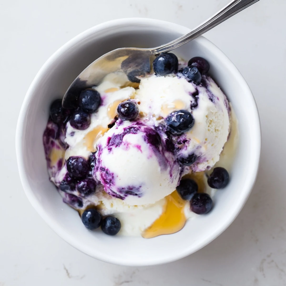A scoop of Blueberry Cottage Cheese Ice Cream in a white bowl, topped with fresh blueberries and a drizzle of honey.
